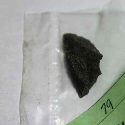 Close up of basalt flake: A piece of dark rock is sealed in a clear plastic bag, placed next to a green label with handwritten notes and the number "363" visible.