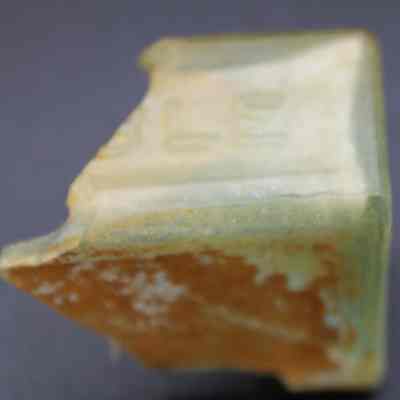 Closeup of glass bottle fragment: The image shows a weathered, greenish bottle fragment with visible edges and embossed letters partially showing "LE", suggesting a piece of historical glassware.
