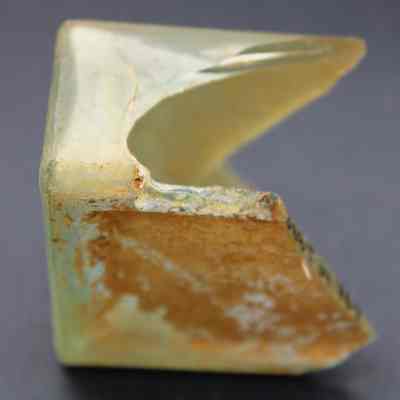Close up of a glass bottle fragment: A triangular-shaped fragment of a glass bottle lies on a dark surface, its edges appearing rough and weathered with hints of soil.