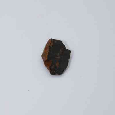 Closeup of basaltic glass flake: A small, dark basaltic glass flake is lying on a plain white surface, displaying its rough edges and natural texture.