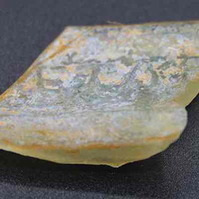 Closeup of glass bottle fragment: The image shows a worn, light-colored glass fragment with rough edges and a slightly curved shape, resting on a dark background.