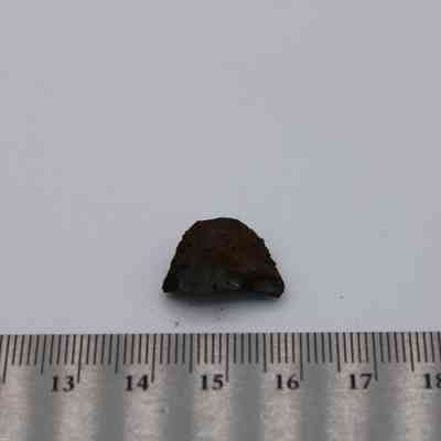 Close up of basaltic glass flake and metal ruler: The image shows a small piece of basaltic glass placed next to a ruler for size reference on a white surface.