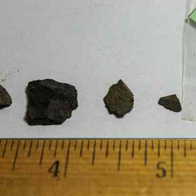 Basalt flakes: Several rock fragments of varying sizes are arranged on a white surface next to a ruler for scale with a plastic bag and green paper.