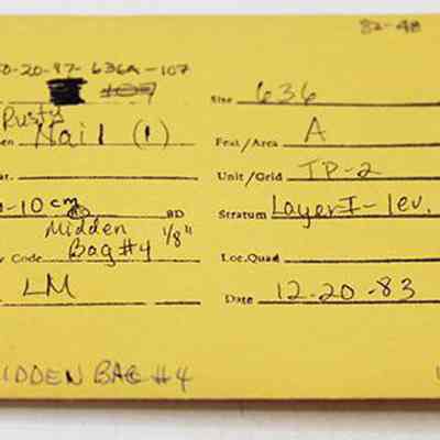 Artifact envelope for rusty nail: The image shows a yellow envelope with handwritten and typed archaeological details, including site, specimen, depth, unit, stratum, recovery code, and date.