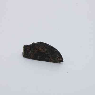 Closeup of basaltic glass flake: The image depicts a small, dark basaltic glass flake with uneven, rough edges resting on a plain white surface.