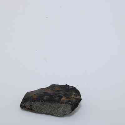 Closeup profile of basaltic glass flake: A small, dark basaltic glass flake rests on a plain white background, showcasing its rough, uneven texture and irregular shape.