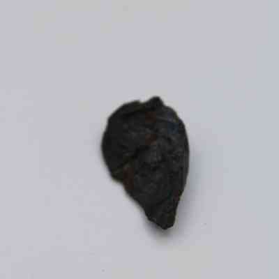 Lengthwise view of triangular basaltic glass flake: The image displays a dark, triangularly-shaped basaltic glass flake with a rough texture, positioned on a plain white background.