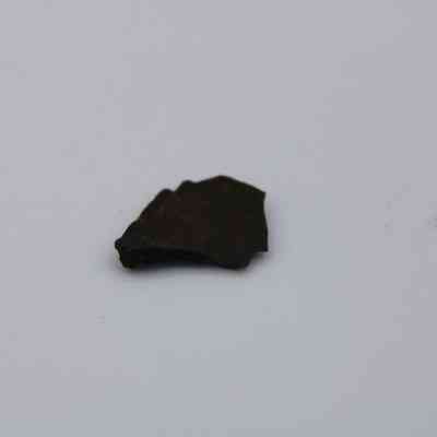 Closeup of basaltic glass flake: The image shows a small, irregularly shaped basaltic glass flake lying on a plain, light surface, highlighting its dark color and texture.