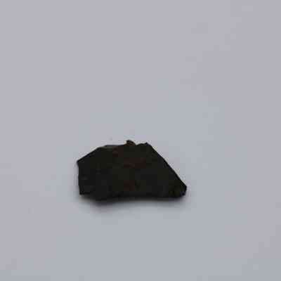 Closeup of basaltic glass flake: The image shows a small, irregularly shaped basaltic glass flake, resting on a plain, light-colored surface.