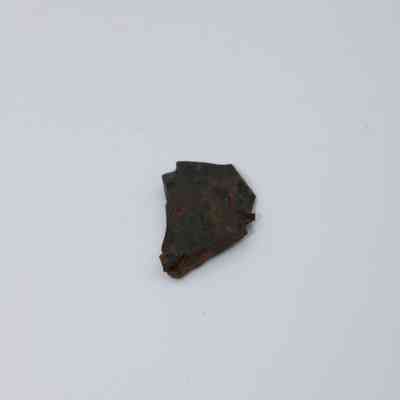Closeup of tiny basaltic glass flake: A small, irregularly-shaped piece of dark basaltic glass flake lies on a plain, light-colored background, showcasing its rough texture.