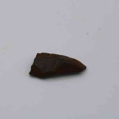 Basaltic glass flake: A small, dark brown basaltic glass flake rests on a plain white surface, its rough edges and slightly glossy texture catching the light.