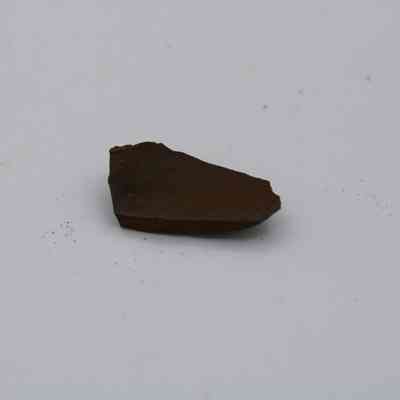 Closeup of basaltic glass flake: A small piece of basaltic glass flake lies on a plain white background, displaying its dark brown color and irregular shape.