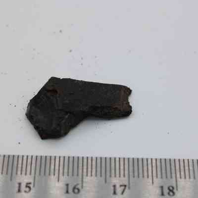Closeup of basaltic glass flake with ruler: The image shows a small basaltic glass flake with a rough texture, lying next to a ruler for scale on a white surface.