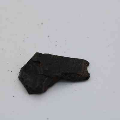 Closeup of basaltic glass flake: A small, dark basaltic glass flake rests on a white surface, showcasing its rough, jagged texture and irregular shape.