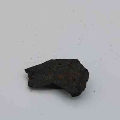 Closeup of basaltic glass flake: A small, dark basaltic glass flake rests on a light surface, showcasing its rough texture and irregular shape, with specks scattered nearby.