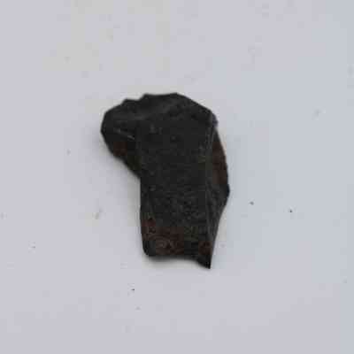 Closeup of lengthwise view of basaltic glass flake: A piece of basaltic glass flake, dark and irregularly shaped, rests against a plain, light-colored background, highlighting its rough texture.