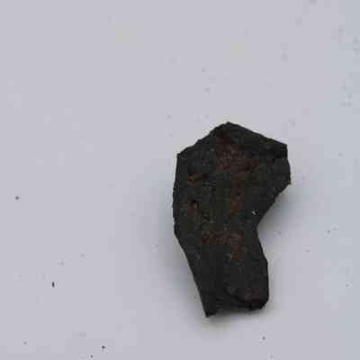Basaltic glass flake: A solitary basaltic glass flake rests on a plain, light background, displaying its dark, rough texture and uneven edges.