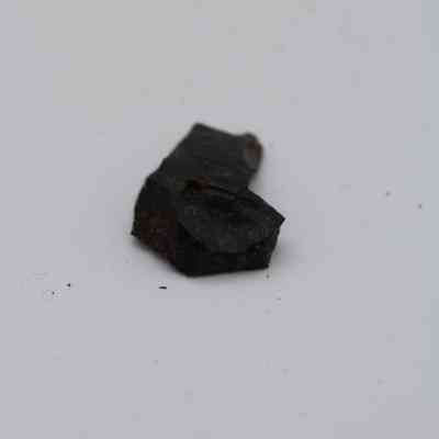 Basaltic glass flake: A small basaltic glass flake rests on a plain white surface, showcasing its dark, glossy appearance and rough texture.