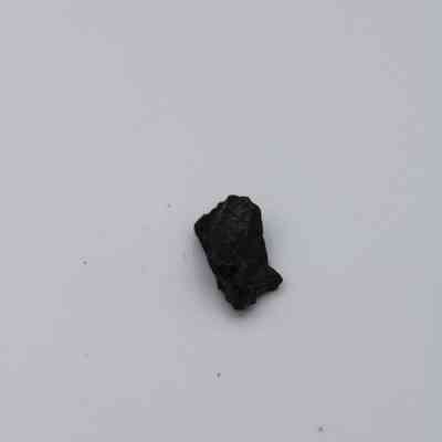 Tiny basaltic glass flake: A single piece of basaltic glass flake rests on a plain, neutral background, highlighting its dark, irregular surface texture.