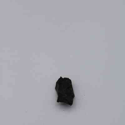 Tiny basaltic glass flake: A dark basaltic glass flake rests on a light, plain surface, displaying rough texture and irregular shape.
