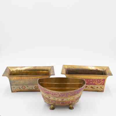 Rectangular cachepots and dhow-shaped cachepot: The image shows two brass rectangular cachepots with Arabic script, one pink and another blue, alongside a purple, dhow-shaped pot.