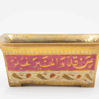 Rectangular cachepot: The image shows a brass cachepot with Arabic script on a pink band, displaying intricate designs underneath. It reads, "whoever offers good deeds..."