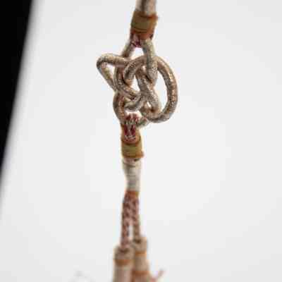 End of corded ropes attached to the clasp of the necklace.: Up close image of the intricate knot on the bottom of the corded clasp ropes. It is in a similar style of Celtic knots and is silver in color. There is a tassel at the end of each cord below the knot.