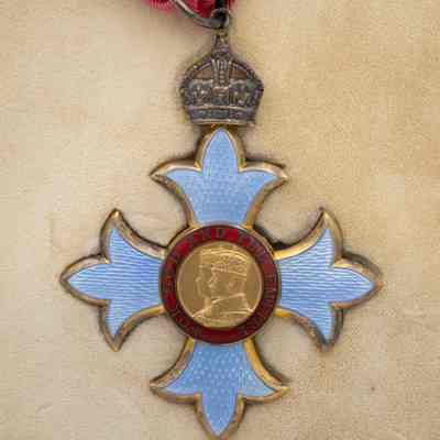 Commander of the British Empire medal: The image shows a Commander of the British Empire medal, featuring a central portrait, surrounded by a red band inscribed with "FOR GOD AND THE EMPIRE." The pale blue Maltese cross is adorned with a crown and suspended from a red and white ribbon.