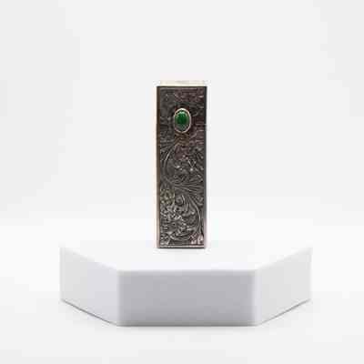 Silver Italian lipstick box gifted to Mrs. Jessie Jordan by the queen consort of Iran.: Image of the front of the silver Italian lipstick box on a stand. The box is intricately decorated with floral vine designs and a green jade stone.