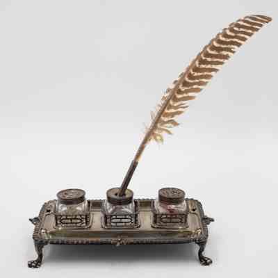 Full ink stand, ink wells, and quill.: The silver ink stand has three ink holders with tags inside, and a striped feather quill in the central ink well.