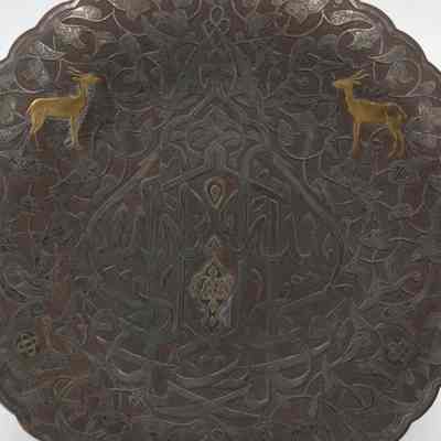 Silver and brass plate inscribed with "Bieznella," or "Welcome" in Arabic.: Up close image of the propped up silver and brass plate. The center of the plate is inscribed with Arabic script reading "Bieznella." There are two small brass deer on the top right and left of the Arabic lettering.