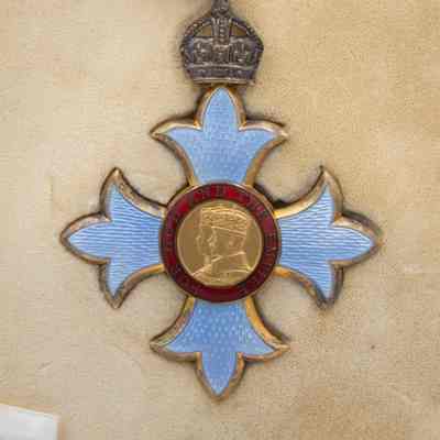 Commander of the British Empire medal: The image shows a Commander of the British Empire medal, featuring a central portrait, surrounded by a red band inscribed with "FOR GOD AND THE EMPIRE." The pale blue Maltese cross is adorned with a crown and suspended from a red and white ribbon.