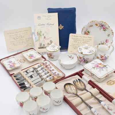 Complete collection of Royal Crown Derby porcelain given to Mrs. Jessie Jordan by the Masonic Lodges.: Image of the full set of Royal Crown Derby items received by Mrs. Jessie Jordan. There is a blue Masonic Presentation Bible propped up in the back along with three invitations from various Ladies' Nights. All of the porcelain items are white and have the same floral pattern decorating them. There is one larger bowl, a cream pitcher, two sugar dishes (one large, one small), a smaller bowl, two cigarette ashtrays, a cigarette box, a set of six silver knives in a red case, a pair of silver salad serving spoons in a red case, and six small teacups.