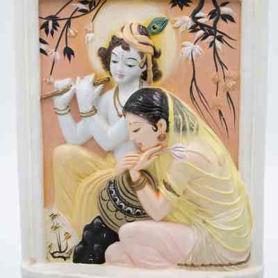 Molded ceramic picture painted with Radha and Lord Krishna, likely from India.: Up close image of the molded ceramic picture painted with Radha and Lord Krishna on a black picture stand. Radha is depicted in light pink clothing with a yellow shawl draped over her head. Krishna is seen behind Radha and is playing a flute with a peacock feather in his hair.