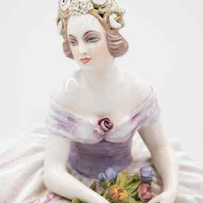 Woman figurine, close-up: A porcelain lady figurine sits elegantly, adorned in blue and lavender, holding colorful flowers.