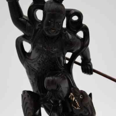 Carved ebony figurine of man with a spear fighting a sea monster.: Image of a dark brown carved figurine of man holding spear and standing on some sort of sea monster. Both the creature and man have white ivory eyes and teeth.
