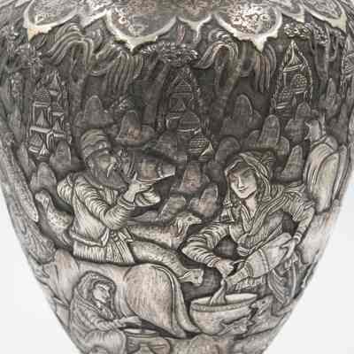 Scene on the silver and brass urn from Persia.: Up close image of the debossed scene on the silver urn. The scene depicts a man drinking from an urn and two women processing milk. One is milking a cow while the other is pouring something into a bowl.