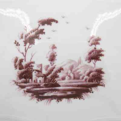 Pastoral scene: The image shows a white ceramic with a burgundy pastoral scene painted on. It has trees, hills, and houses.