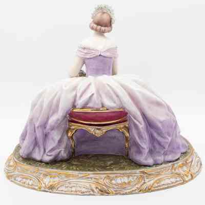 Back of figurine: A porcelain lady figurine in blue and lavender sits elegantly on a rose pillow with a gold base.