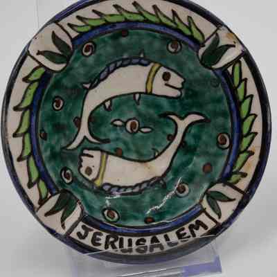One of the four painted ceramic ashtrays given to the Jordans by a ruler in Bombay.: Up close image of one of the four painted ceramic ashtrays. This one is painted with a blue background and two white fish circling in the center. Around the outside there are green leaves and it reads "JERUSALEM" at the bottom in black text.