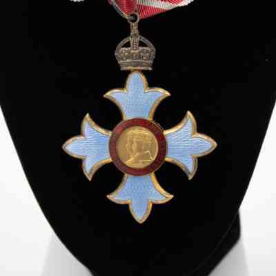 Commander of the British Empire medal: The image shows a Commander of the British Empire medal, featuring a central portrait, surrounded by a red band inscribed with "FOR GOD AND THE EMPIRE." The pale blue Maltese cross is adorned with a crown and suspended from a red and white ribbon.
