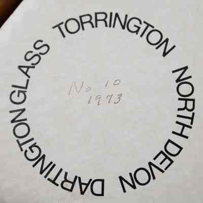 Bottom of the triangular box that the Dartington Glass commemorative tankard came in.: Up close image of the bottom of the triangular cardboard box. The bottom is white with black printed text that reads "DARTING GLASS TORRINGTON NORTH DEVON" and black handwriting that reads "No 10 1873."