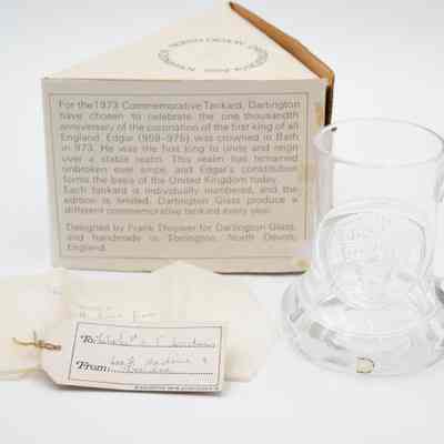 Side of triangle box, Dartington Glass tankard, and notes.: Image of the glass tankard pressed with King Edgar's head. There is a small sticker on the bottom that says "Dartington England." Two notes found with the tankard. The card reads "To: Mr. + Mrs. L.T. Jordan" and "From: Jack, Nadine, + Deirdre." The tissue paper reads "To Jessie with love from Deirdre." Back of the triangular box has the story of King Edgar's coronation: "For the 1873 Commemorative Tankard, Dartington have chosen to celebrate the one thousandth anniversary of the coronation of the first king of all England. Edgar (959-975) was crowned in Bath in 973. He was the first king to unite and reign over a stable realm. This realm has remained unbroken ever since, and Edgar's constitution forms the basis of the United Kingdom today. Each tankard is individually numbered, and the edition is limited. Dartington Glass produce a different commemorative tankard every year. Designed by Frank Thrower for Dartington Glass, and handmade in Torrington, North Devon, England."