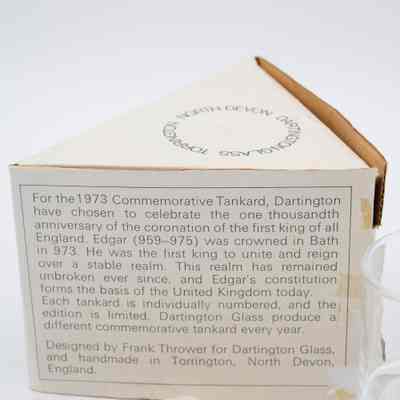 Description of the tankard on one of the sides of the triangular box.: Up close image of the description on the triangular box. It has the story of King Edgar's coronation: "For the 1873 Commemorative Tankard, Dartington have chosen to celebrate the one thousandth anniversary of the coronation of the first king of all England. Edgar (959-975) was crowned in Bath in 973. He was the first king to unite and reign over a stable realm. This realm has remained unbroken ever since, and Edgar's constitution forms the basis of the United Kingdom today. Each tankard is individually numbered, and the edition is limited. Dartington Glass produce a different commemorative tankard every year. Designed by Frank Thrower for Dartington Glass, and handmade in Torrington, North Devon, England."