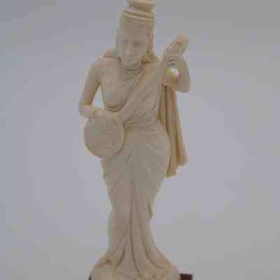 Figure of woman holding stringed instrument: The image depicts an intricately carved ivory figurine of a woman holding a stringed instrument. She wears a long layered dress fitted to her body. She also has a tall hat on.