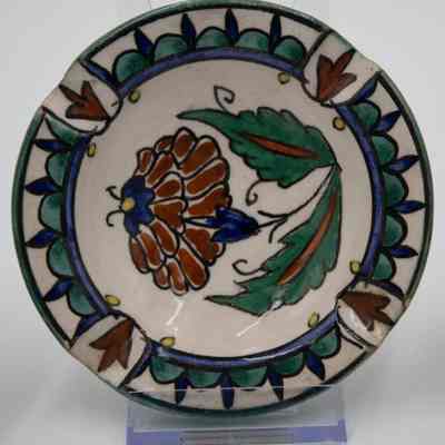 One of the four painted ceramic ashtrays given to the Jordans by a ruler in Bombay.: Up close image of one of the four painted ceramic ashtrays. This one is painted with a white background and brown and green flower in the center. Around the outside there are blue and teal decorations.