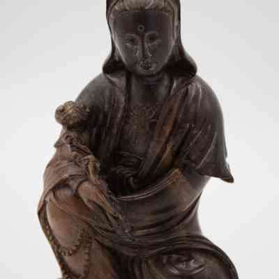 Carved wooden figurine of a seated woman from Bali.: Up close image of the carved wooden figurine of a seated woman. It depicts the Chinese goddess of mercy, Guanyin, in her traditional "royal-ease" pose. She is wearing a long robe and holding a flower.