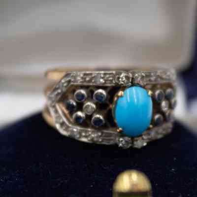 Gold-banded ring with small diamonds and a turquoise stone set in silver.: Up close image of the gold-banded ring with six small stones on either side of the large turquoise stone in a pronged-setting in the middle. There is silver detail around the edges of the small stone and it is in a blue corduroy ring box. The ring is sitting on blue felt.
