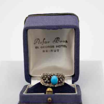 Gold-banded ring with small diamonds and a turquoise stone set in silver.: Up close image of the gold-banded ring with six small stones on either side of the large turquoise stone in a pronged-setting in the middle. There is silver detail around the edges of the small stone and it is in a blue corduroy ring box. The ring is sitting on blue felt and the inside of the lid reads "Asfar Bros ST. GEORGE HOTEL BEIRUT" in black writing on white satin.