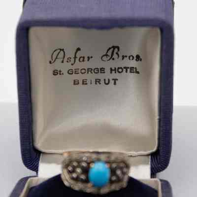 Inside of the blue corduroy ring box.: Up close image of the inside of the lid, it reads "Asfar Bros ST. GEORGE HOTEL BEIRUT" in black writing on white satin.